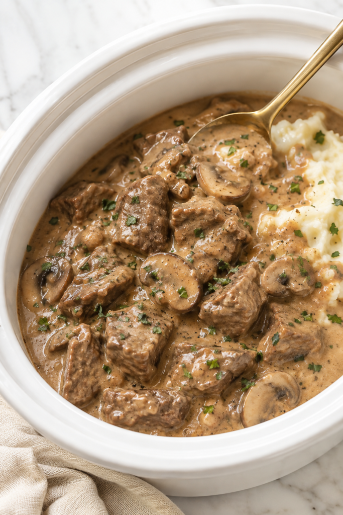 Tender slow cooker creamy beef tips with mushrooms and rich gravy served beside mashed potatoes in a white Crockpot.