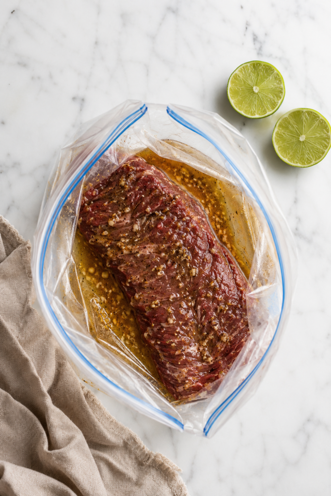 Raw skirt steak marinating with lime, garlic, cumin, and soy sauce in an open zip-top bag.