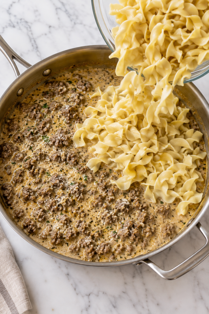 Cooked egg noodles being poured into creamy cowboy butter ground beef sauce in a large skillet before tossing
