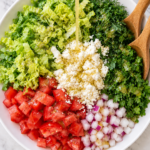 Lemon vinaigrette being poured over separate piles of romaine, tomatoes, red onion, fresh dill, parsley, and feta in a white bowl with wooden salad tossers.