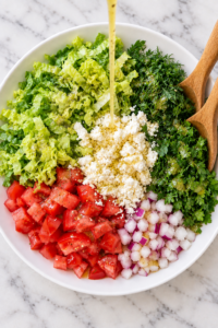 Lemon vinaigrette being poured over separate piles of romaine, tomatoes, red onion, fresh dill, parsley, and feta in a white bowl with wooden salad tossers.
