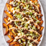 Big Mac inspired poutine with crispy fries, ground beef, cheese curds, pickles, lettuce, onions, and special sauce on a white oval platter