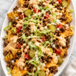 Loaded Big Mac nachos with seasoned ground beef, melted cheese, pickles, lettuce, tomatoes, and creamy burger sauce on a large white oval platter.
