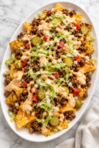 Loaded Big Mac nachos with seasoned ground beef, melted cheese, pickles, lettuce, tomatoes, and creamy burger sauce on a large white oval platter.