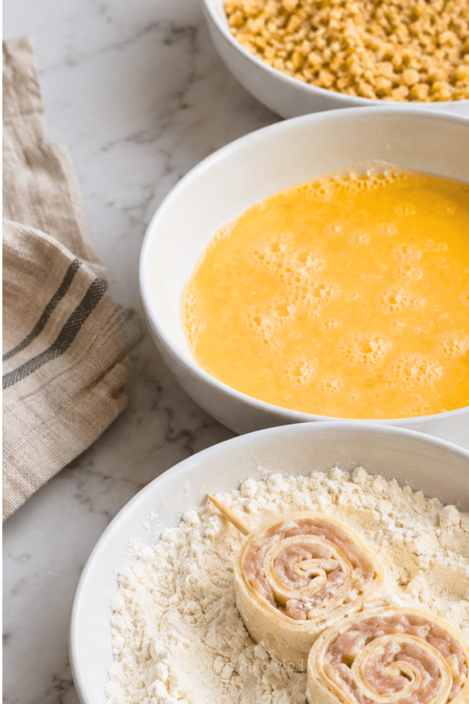 Raw ground chicken pinwheel skewers coated in flour beside bowls of egg wash and seasoned panko breadcrumbs.