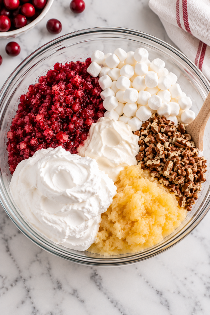 Macerated cranberries, pineapple, mini marshmallows, pecans, sour cream, and whipped topping in a bowl before mixing cranberry marshmallow salad.