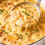 Silver ladle scooping creamy copycat Outback Walkabout onion soup with caramelized onions and melted cheese from a white enamel pot