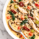 Creamy Tuscan chicken wild rice soup with shredded chicken, spinach, sun-dried tomatoes, Parmesan, carrots, and celery in a white bowl.