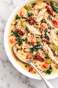 Creamy Tuscan chicken wild rice soup with shredded chicken, spinach, sun-dried tomatoes, Parmesan, carrots, and celery in a white bowl.