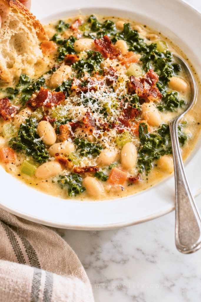 Bread dipping into creamy Tuscan white bean and kale soup with bacon in a white bowl.
