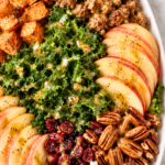 Crispy sweet potato and sausage harvest salad with kale, apples, cranberries, pecans, goat cheese, and maple Dijon dressing on a white platter.