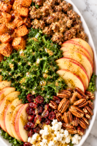 Crispy sweet potato and sausage harvest salad with kale, apples, cranberries, pecans, goat cheese, and maple Dijon dressing on a white platter.