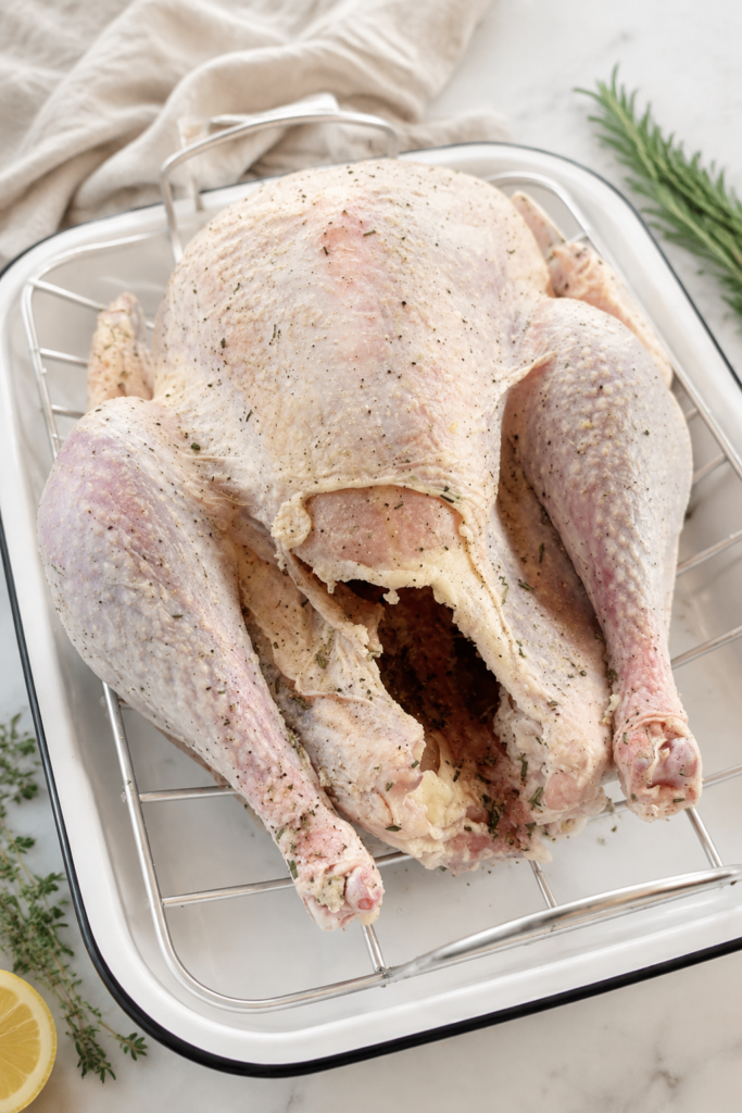 Raw whole turkey covered in dry brine seasoning on a roasting rack inside a white enamel roasting pan.