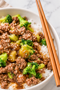 Ground beef and broccoli over white rice with brown sauce and sesame seeds in a white bowl with wooden chopsticks.