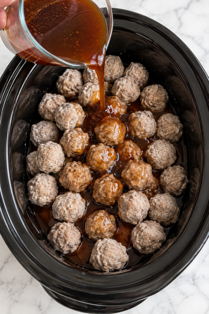 Apple cider sauce being poured over frozen meatballs in a black slow cooker.