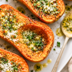 Roasted butternut squash halves drizzled with garlic cowboy butter, fresh herbs, and Parmesan on a white platter.