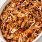 Tender slow cooker apple cider pulled pork with caramelized onions and barbecue sauce in a white oval serving dish.