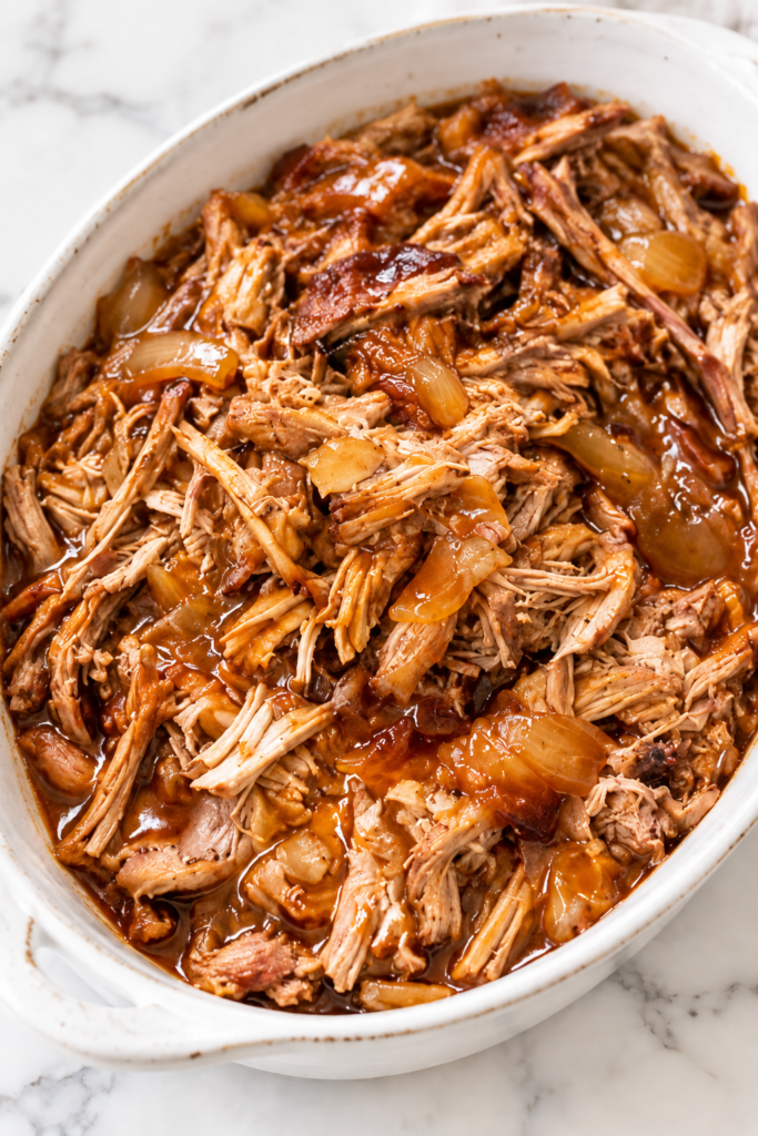 Tender slow cooker apple cider pulled pork with caramelized onions and barbecue sauce in a white oval serving dish.
