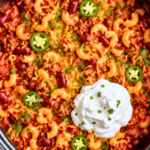 heesy slow cooker chili mac with ground beef, kidney beans, elbow macaroni, jalapeños, cilantro, and sour cream in a black crockpot.