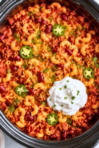 heesy slow cooker chili mac with ground beef, kidney beans, elbow macaroni, jalapeños, cilantro, and sour cream in a black crockpot.