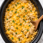 Bird’s-eye view of creamy slow cooker cowboy butter chicken and rice casserole topped with melted cheddar, shredded chicken, parsley, and black pepper in a black crockpot.