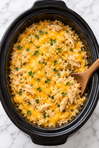 Bird’s-eye view of creamy slow cooker cowboy butter chicken and rice casserole topped with melted cheddar, shredded chicken, parsley, and black pepper in a black crockpot.