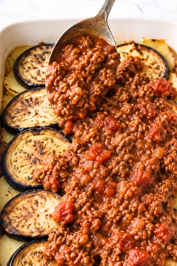 Spoon adding thick meat sauce over roasted eggplant and potatoes in a white baking dish for Greek moussaka.