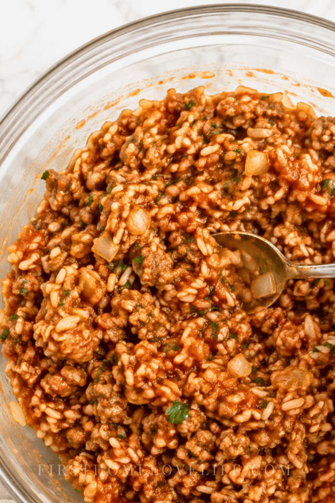 A hand mixes savory ground beef, rice, onion, and tomato filling in a large glass bowl for baked stuffed onions.