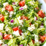 Close-up of tossed Greek salad with romaine ribbons, tomatoes, red onion, dill, parsley, feta, and lemon vinaigrette in a white bowl.