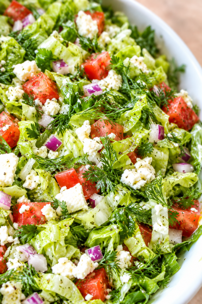 Close-up of tossed Greek salad with romaine ribbons, tomatoes, red onion, dill, parsley, feta, and lemon vinaigrette in a white bowl.