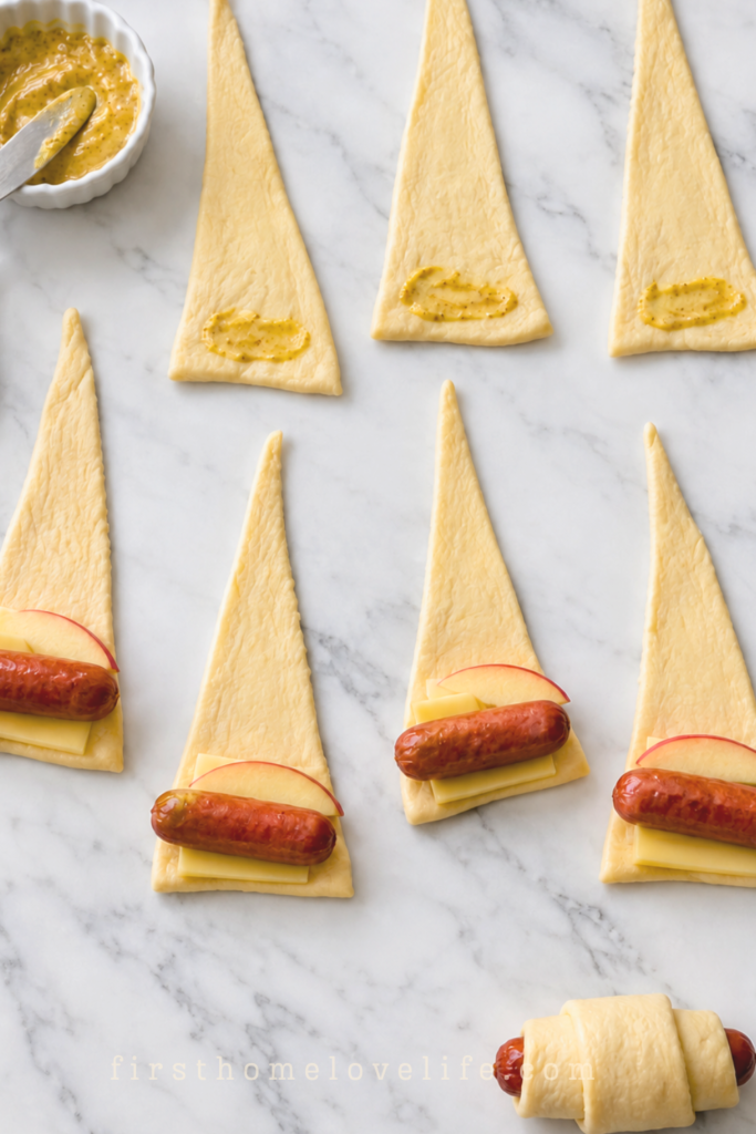 Crescent dough triangles with Dijon mustard, Gouda, apple slices, and cocktail sausages in various stages of assembly on marble.