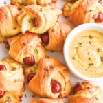 Apple Gouda pigs in a blanket with golden crescent rolls and a light thyme garnish on a white platter beside maple mustard dip.
