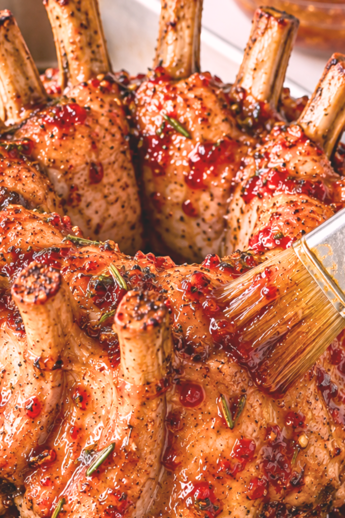 Brushing sweet and spicy red pepper jelly glaze onto a golden-brown pork crown roast in a stainless steel roasting pan.