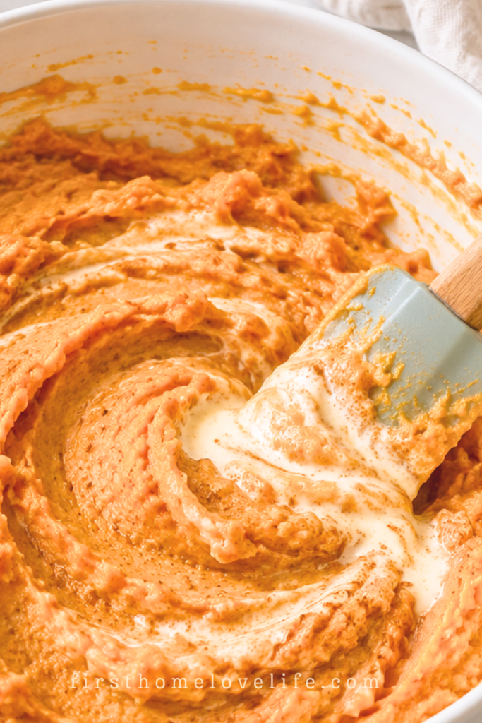 Cream being folded into orange sweet potato casserole filling with a spatula in a large white mixing bowl on marble.