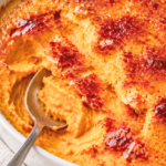 Crème brûlée sweet potato casserole with a cracked caramelized sugar topping and a spoon revealing creamy sweet potatoes in a white baking dish.
