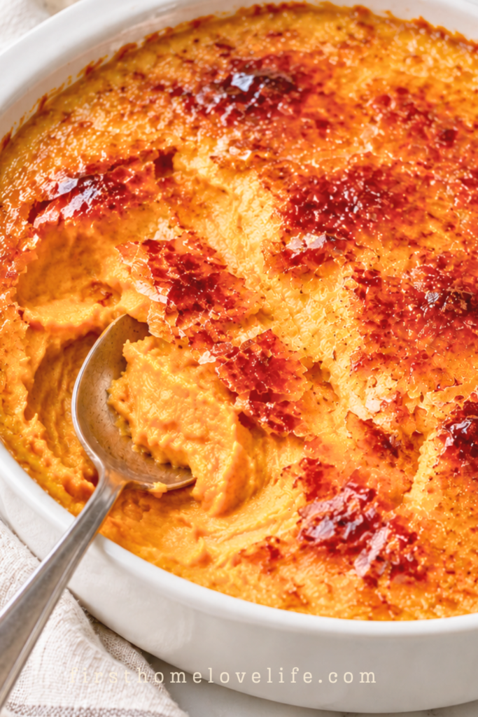Crème brûlée sweet potato casserole with a cracked caramelized sugar topping and a spoon revealing creamy sweet potatoes in a white baking dish.