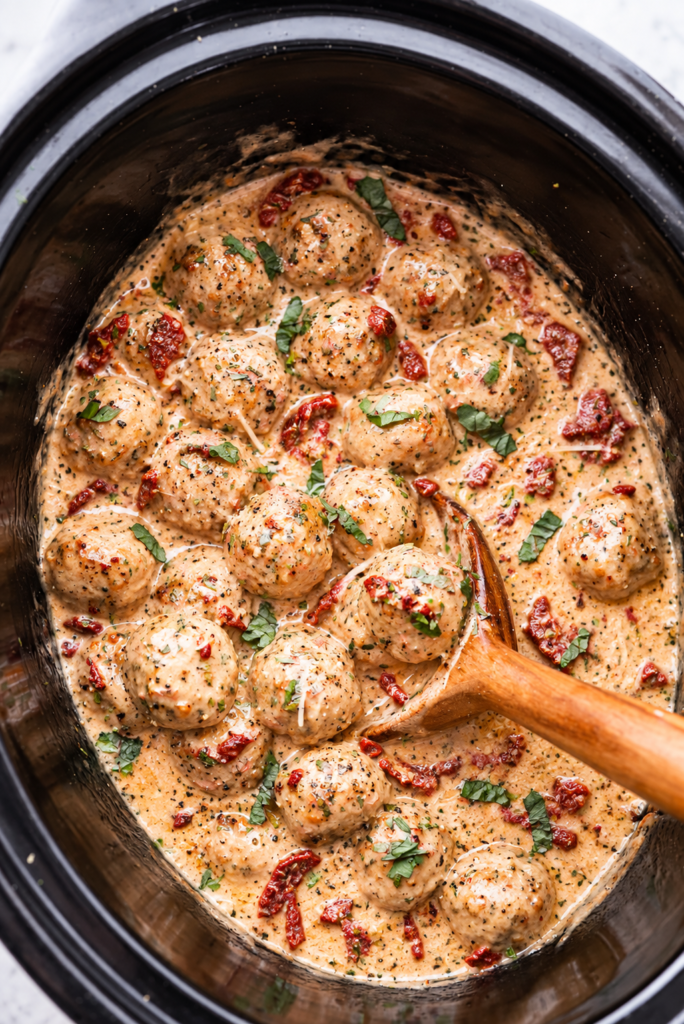 Creamy Crockpot Marry Me Chicken Meatballs with sun-dried tomatoes, parmesan, and fresh basil in a black slow cooker.