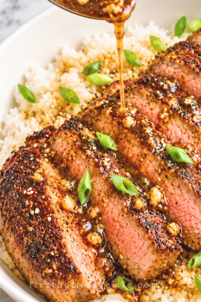 Sliced ginger miso sirloin steak over white rice with green onions and sesame seeds, topped with garlic butter sauce drizzled from a spoon.