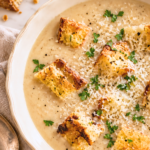 Creamy garlic bread soup topped with golden croutons, grated Parmesan, and parsley in an ivory bowl on a marble counter.