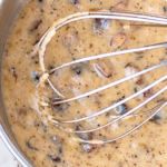 Homemade cream of mushroom soup with mushroom pieces and a wire whisk in a stainless steel pot on a white marble countertop.