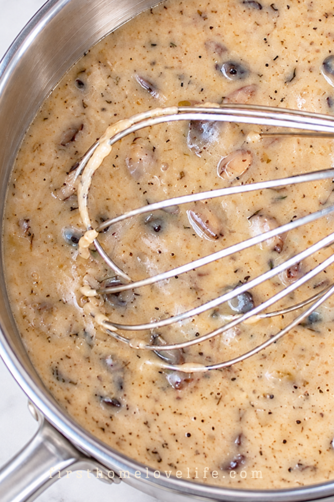 Homemade cream of mushroom soup with mushroom pieces and a wire whisk in a stainless steel pot on a white marble countertop.