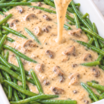 Homemade cream of mushroom soup poured from a silver saucepan over long green beans in a white casserole dish on marble.