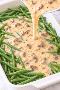 Homemade cream of mushroom soup poured from a silver saucepan over long green beans in a white casserole dish on marble.