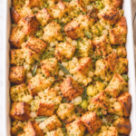 Overhead view of homemade sourdough stuffing with golden toasted bread cubes, celery, onions, and fresh herbs in a white casserole dish.