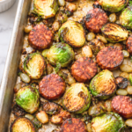 Roasted Brussels sprouts and sliced chorizo on a sheet pan with smoky lime dipping sauce on a white marble counter.