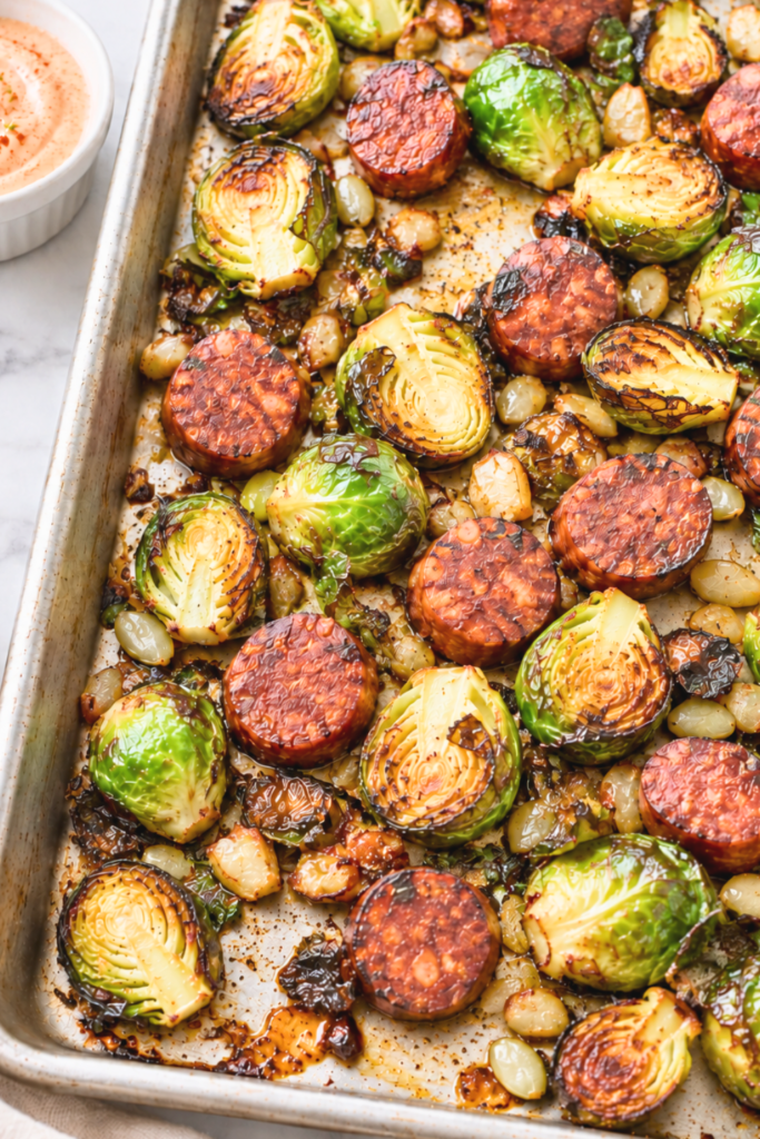 Roasted Brussels sprouts and sliced chorizo on a sheet pan with smoky lime dipping sauce on a white marble counter.