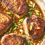 Four pan-seared ginger miso sirloin steaks in garlic butter sauce with green onions and sesame seeds in a stainless steel skillet.