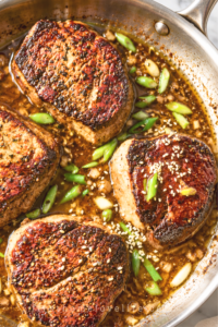 Four pan-seared ginger miso sirloin steaks in garlic butter sauce with green onions and sesame seeds in a stainless steel skillet.