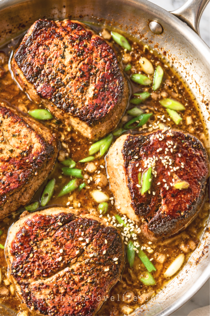 Four pan-seared ginger miso sirloin steaks in garlic butter sauce with green onions and sesame seeds in a stainless steel skillet.