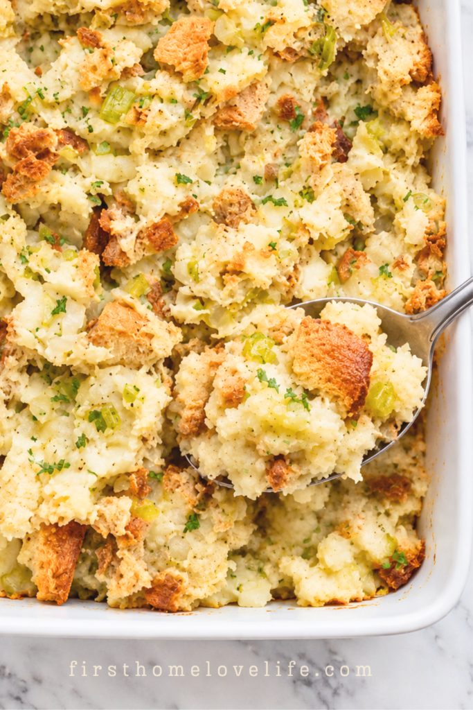 Silver spoon lifting creamy Pennsylvania Dutch potato filling with golden bread pieces, celery, and parsley from a white casserole dish.